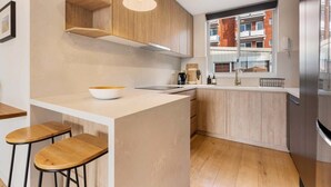 A modern kitchen with wooden cabinets, a central island with stools, a built-in oven, and a window overlooking a residential area. - Coogee Beach Escape | Pool & Balcony (Randwick)