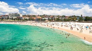 A beach with clear turquoise water, a sandy shore, and a backdrop of buildings. - Coogee Beach Escape | Pool & Balcony (Randwick)