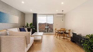 A modern living room with a sofa, a coffee table, and a dining area with chairs. There is a large window with a view of buildings outside. - Coogee Beach Escape | Pool & Balcony (Randwick)
