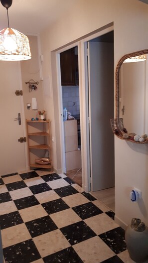 Um corredor com piso xadrez, uma porta que dá para um banheiro, uma prateleira com produtos de higiene pessoal e um lustre.