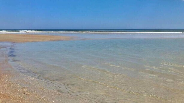 Une plage à l’eau claire et au rivage sablonneux, sous un ciel bleu parsemé de nuages.