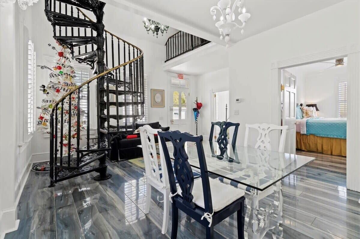 A spacious room with a glass dining table, white chairs, a black spiral staircase, and a chandelier.