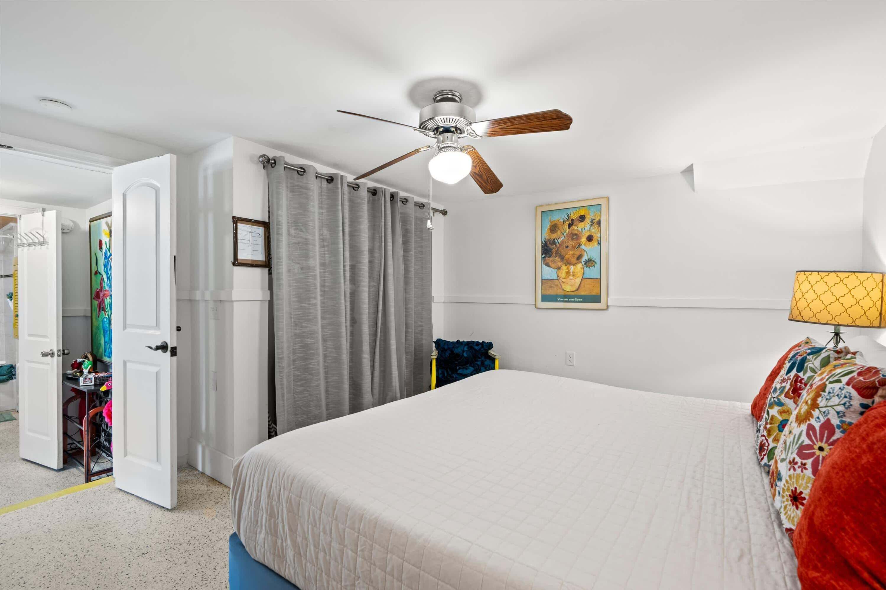 A bedroom with a bed, a ceiling fan, a window with curtains, and a painting on the wall.