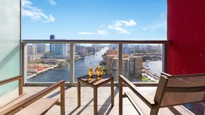 Een balkon met uitzicht op een rivier en stadsbeeld, met houten meubels en twee flessen op een bijzettafeltje.