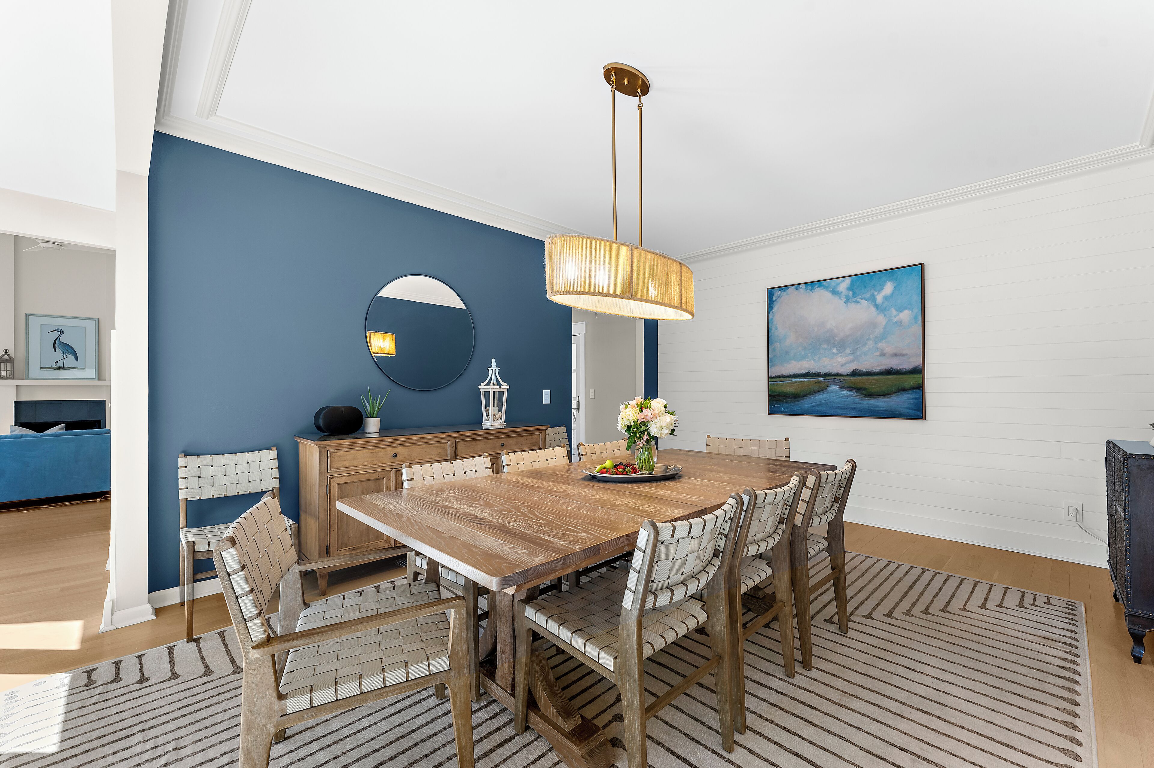 A dining area with a wooden table, white cushioned chairs, a hanging light fixture, a round mirror, and a landscape painting on the wall.