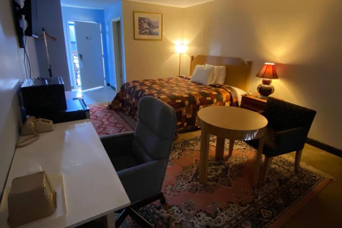 A hotel room with a bed, a desk, a chair, a table, and a lamp.