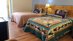 A room with two beds, one with a patterned quilt and the other with a plain bedspread. There is a wooden headboard, a bedside table with a lamp, and a window with curtains. - Lake Country Inn  (Trinity)