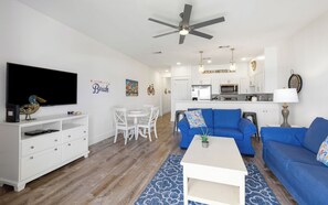 A modern living room with a flat-screen TV, white entertainment unit, blue sofas, and a dining area with white chairs.