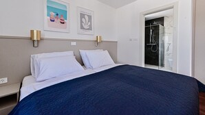 A hotel room with a large bed, two wall-mounted lamps, a framed artwork, and a glass-enclosed shower area.
