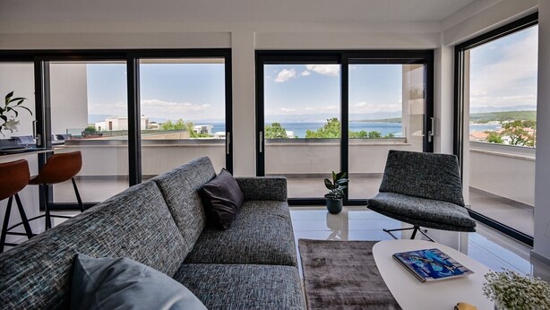 Ein modernes Wohnzimmer mit einer grauen Couch, einem kleinen Tisch und einem Sessel. Große Fenster bieten Blick auf das Meer und die Stadt.
