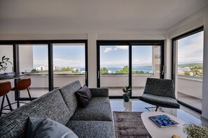 A modern living room with a grey sofa, a small table, and a chair. There are large windows with a view of the sea and buildings. - Olive garden apartment with private pool (Malinska)