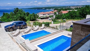 Ein Poolbereich auf dem Dach mit zwei Schwimmbecken, Liegestühlen und Blick auf das Meer sowie Gebäude im Hintergrund.