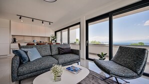 Ein modernes Wohnzimmer mit einem grauen Sofa, einem passenden Sessel und einem Couchtisch. Im Hintergrund befindet sich ein Küchenbereich mit Schränken und einem Esstisch.