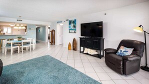 Una sala de estar con televisor, un sillón de cuero, un tapete azul y un comedor con mesa y sillas.