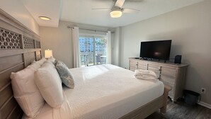 Una cama bien hecha con sábanas blancas y almohadones decorativos, un televisor en una cómoda y un balcón con vistas a edificios y palmeras.