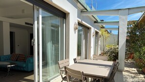 Eine Außenpatio mit Tisch und Stühlen, eine Glasschiebetür, die zu einem Zimmer führt, und ein Gartenbereich mit Pflanzen.