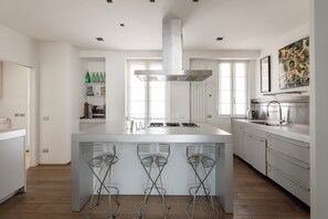 A modern kitchen with a central island, bar stools, a stainless steel range hood, and a window with blinds.