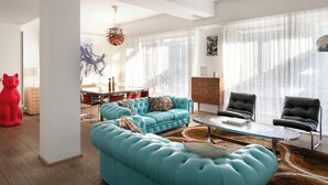 Ein modernes Wohnzimmer mit einem türkisfarbenen Sofa, schwarzen Sesseln, einem Glastisch und einem Essbereich im Hintergrund.