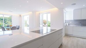 A modern kitchen with white cabinetry, a large island, and a view of greenery outside. - Spacious villa with pool, hot tub and large garden (Montinhos Da Luz)