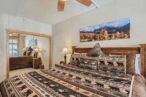 Quarto com uma cama grande, um espelho, um quadro de uma paisagem de montanhas e uma janela com persianas.