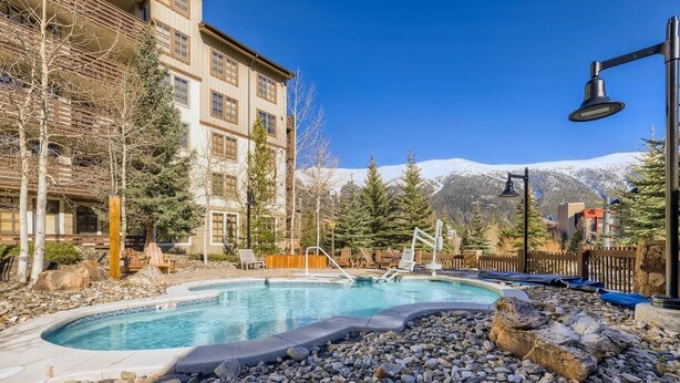 Zona de piscina con jacuzzi, rodeada de terreno rocoso y árboles, con montañas al fondo.