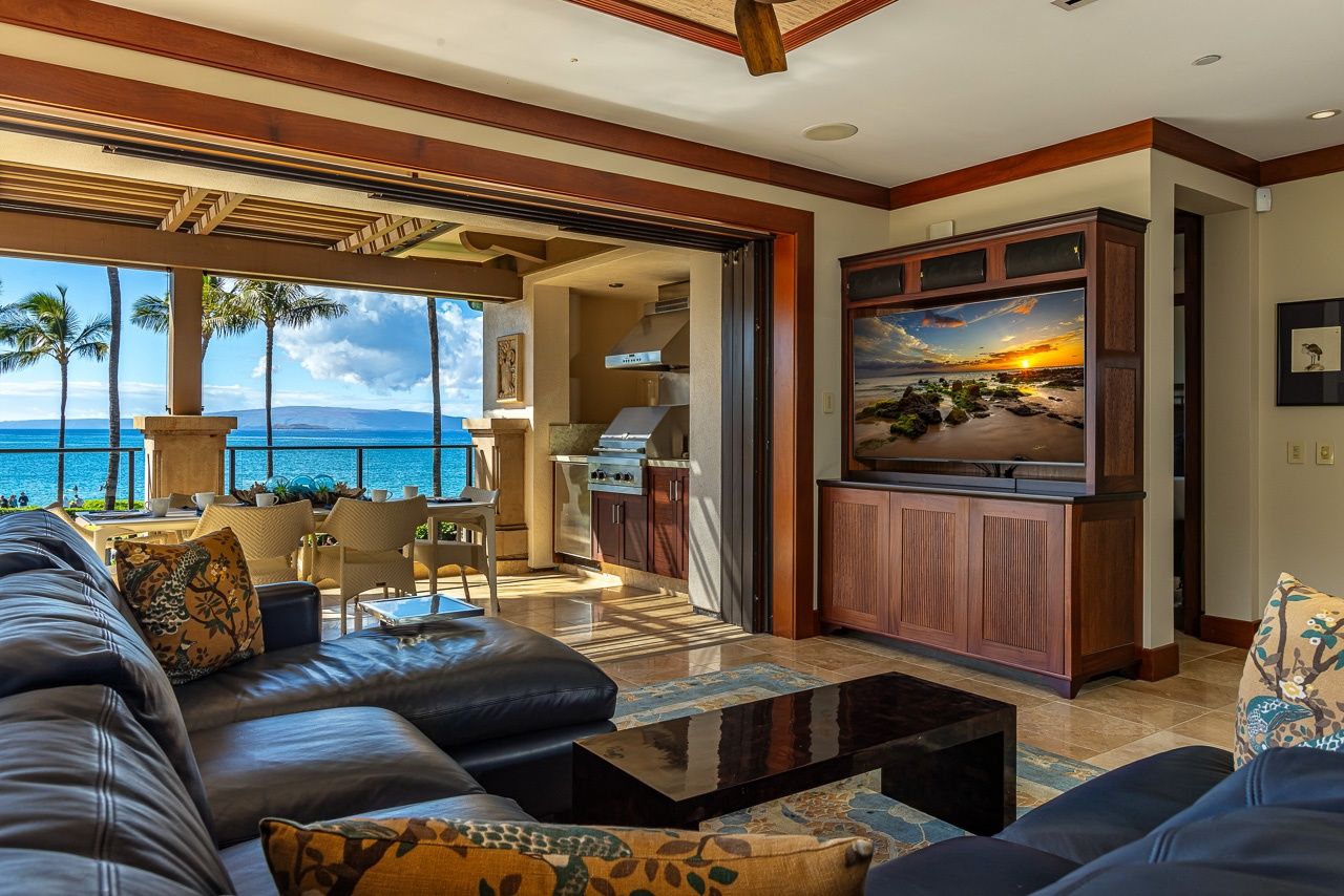 A spacious living room with a large black leather sofa, a flat-screen TV mounted on a wooden cabinet, and a view of the ocean through large windows.