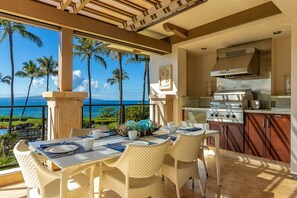 A dining area with a view of the ocean, a kitchen with a stove, and a dining table set for four. - Wailea Beach Villa B202 3 Bedroom Condo (Kihei)