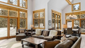 A spacious living room with large windows, a leather sofa set, a wooden coffee table, and a dining area with chairs and a chandelier.