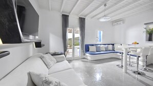 Een moderne woonkamer met een witte bank, een blauwe accentbank, een eethoek met witte stoelen, en een grote televisie aan de muur.