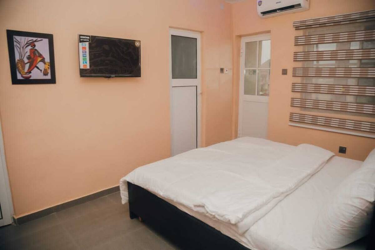 A bedroom with a bed, a door, a window with blinds, and a wall-mounted TV.