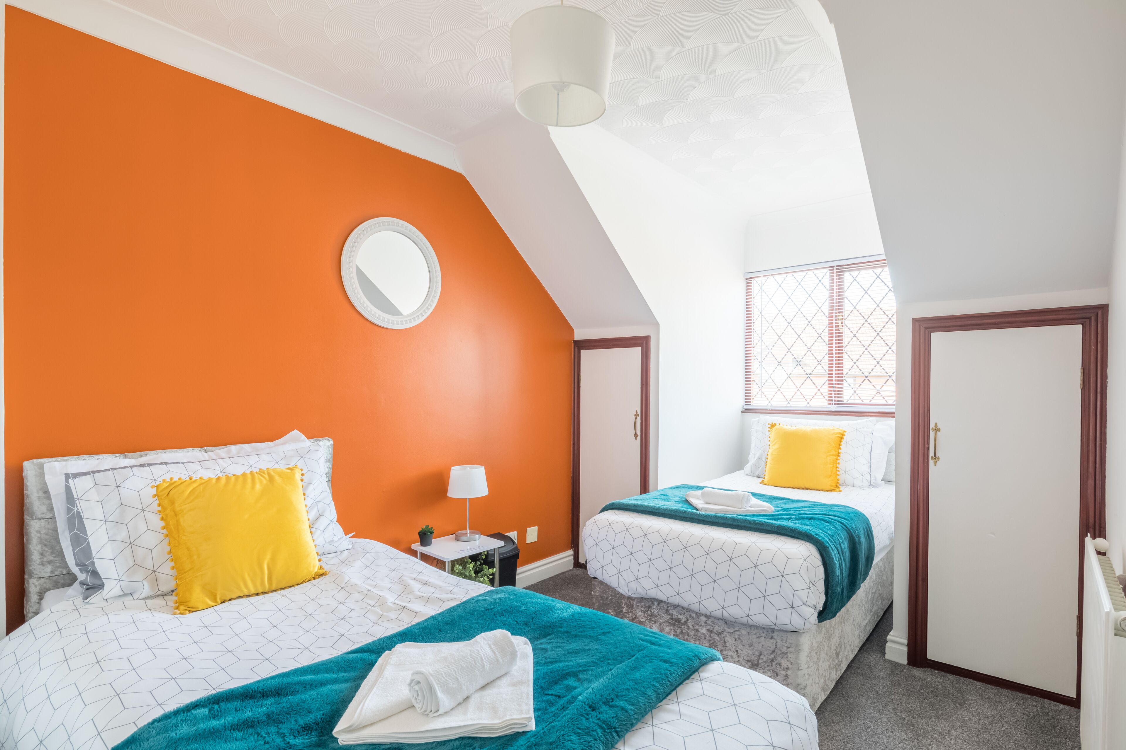 Ein Hotelzimmer mit zwei Betten, orangefarbenen Wänden, einem runden Spiegel und einem Fenster mit Jalousien.