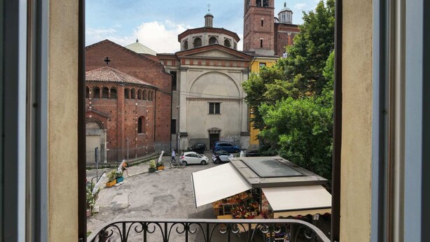 On peut voir une église avec un clocher et un dôme, une rue pavée bordée de voitures garées, et une balustrade de balcon.