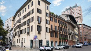 Un immeuble à plusieurs étages, doté d’une façade beige, de nombreuses fenêtres et de balcons. On peut voir des voitures garées devant, et un panneau de signalisation est visible.