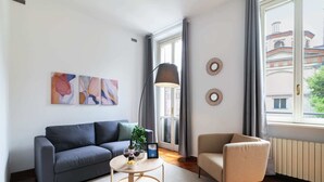 Un salon moderne avec un canapé gris, une table basse ronde et un fauteuil beige. Les murs sont ornés de tableaux abstraits, et on y trouve une lampe sur pied ainsi qu’une fenêtre donnant sur des immeubles.
