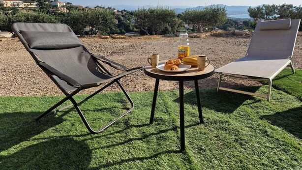 Two lounge chairs, a small round table with food and drinks, and a scenic view of a hillside with buildings and a body of water in the background.