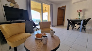 A living room with a TV, two yellow armchairs, a round wooden coffee table with a plate of croissants and two coffee mugs, a dining area with a table and chairs, and a view of the outdoors through a large window.