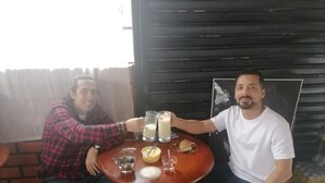 Two people sitting at a round wooden table with a glass of milk, a plate with a cookie, and a metal cup. - Intipunku hospedaje (Cusco)