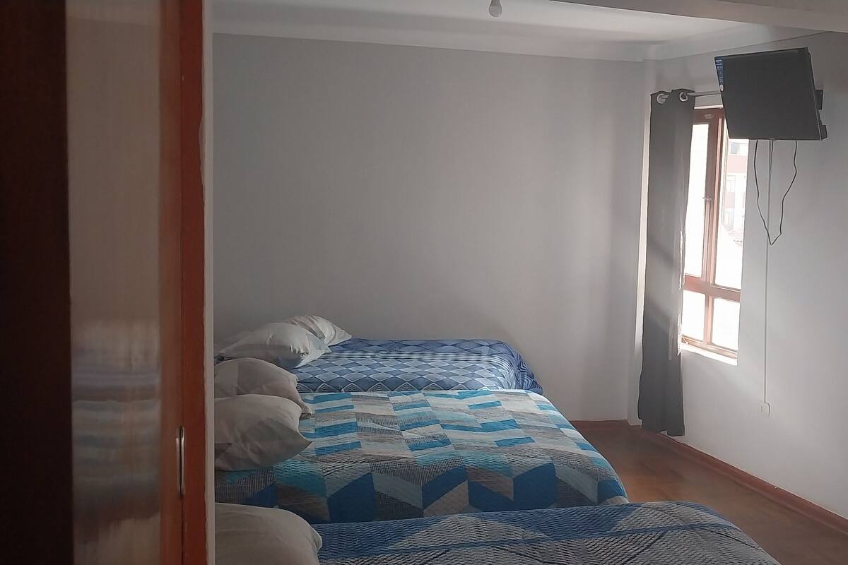 A bedroom with two beds, a TV mounted on the wall, and a window providing natural light.