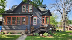 A two-story house with a porch, a chimney, and a garden with plants and a pathway. - Martha's Place (Altura)