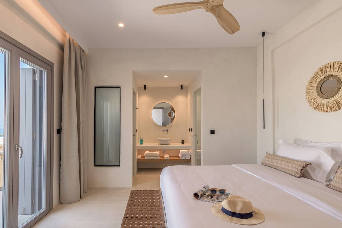 Un dormitorio de hotel moderno con una cama grande, un ventilador de techo, un espejo redondo y vistas a la playa a través de puertas correderas.