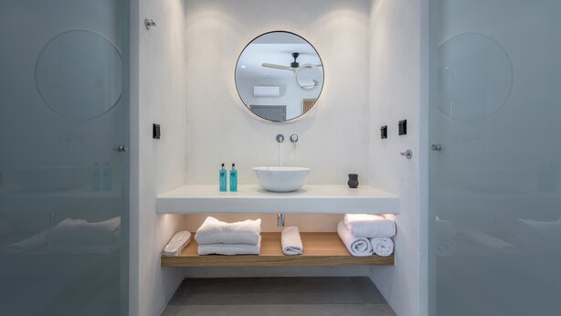 Ein modernes Badezimmer mit einem weißen Waschbecken, einem runden Spiegel und einem hölzernen Ablagebrett mit Handtüchern.
