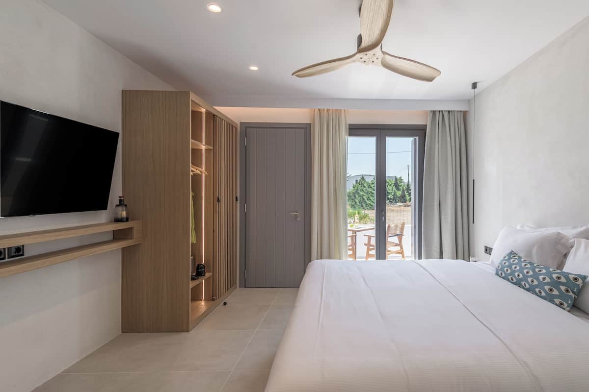 Una habitación de hotel moderna con una cama grande, un televisor montado en la pared, un armario y un balcón con mobiliario exterior.