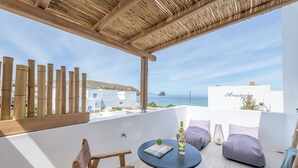 Een balkon met een houten pergola, een rieten stoel, een ronde tafel met een boek en een fles, en paarse kussens.
