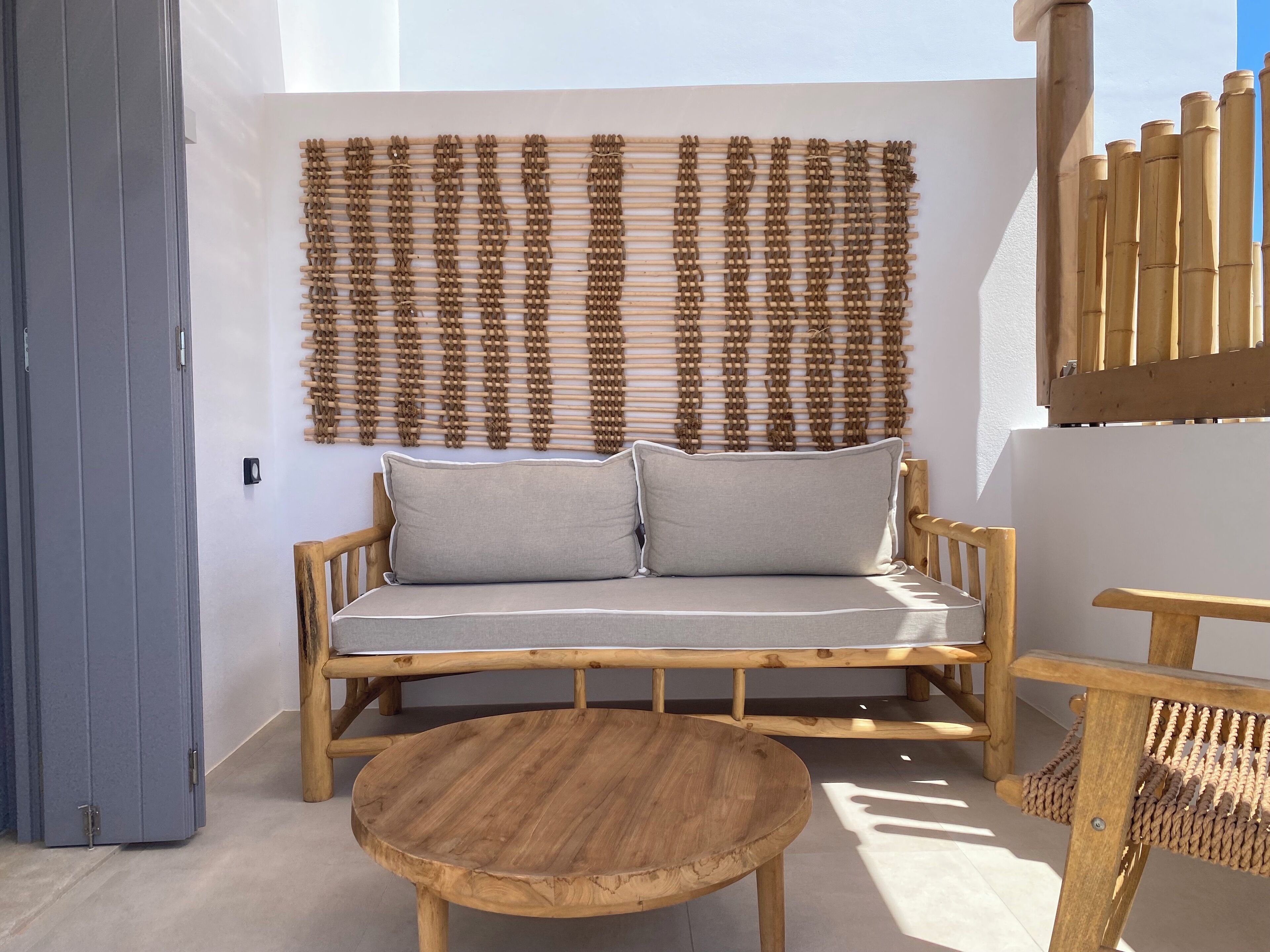 Un canapé en bois avec des coussins, une table ronde en bois et une tapisserie murale tissée dans une pièce lumineuse, dotée d’une porte et d’une rambarde en bois.