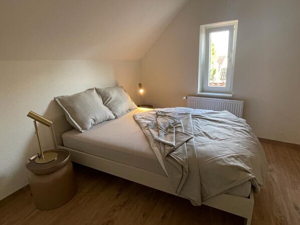 Una cama individual en una habitación con techo inclinado, una mesita de noche pequeña con una lámpara y una ventana que da a zonas verdes.