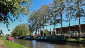 A canal with narrowboats, a grassy path, and a wooden building with a deck. - Chetwynd Lodge (Stafford)