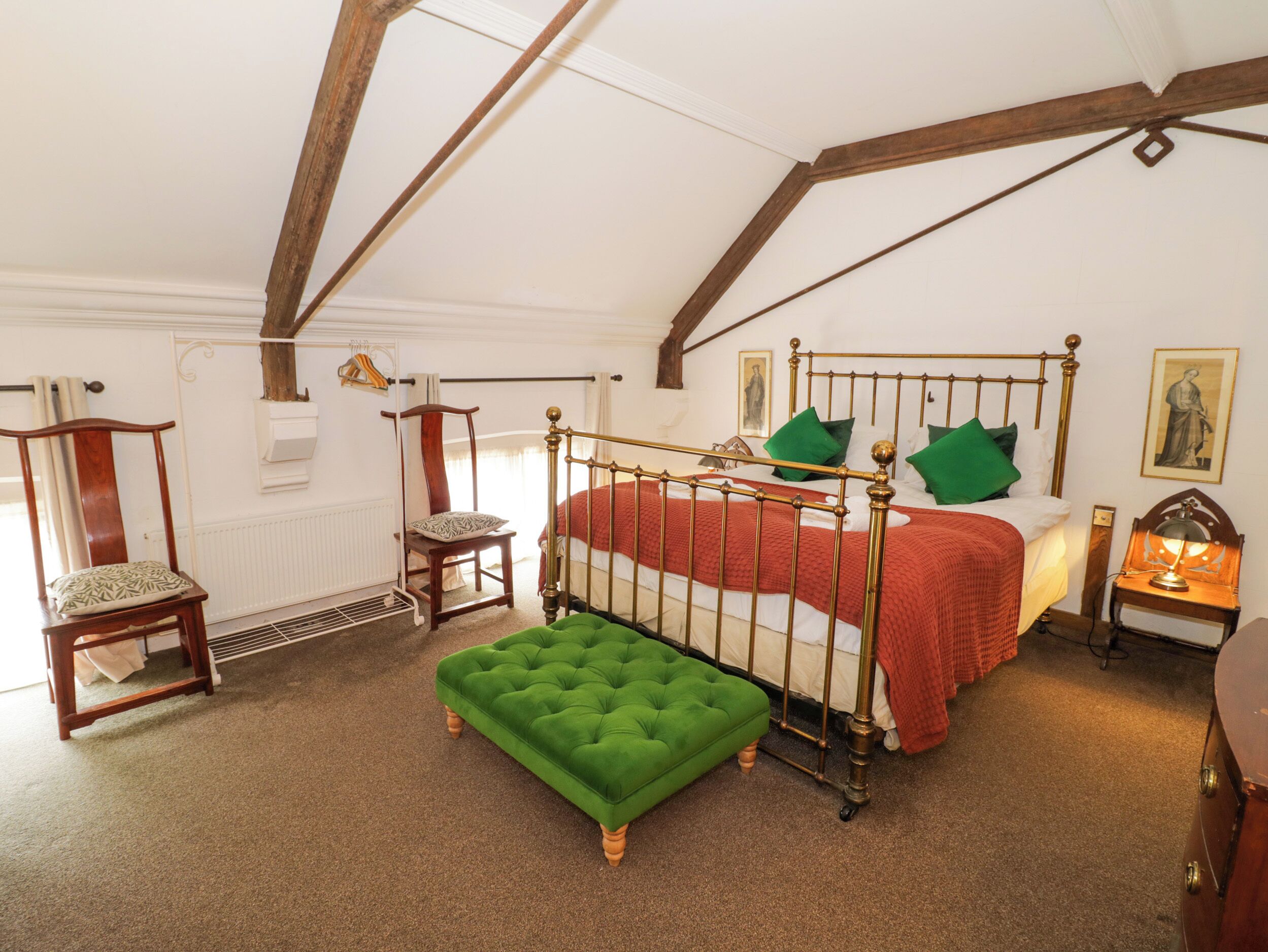 A bedroom with a four-poster bed, a green ottoman, a wooden chair, and a nightstand with a lamp.