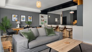 Un salon moderne avec un canapé gris, des coussins verts, une table basse en bois et un coin repas comprenant un buffet blanc et des tabourets de bar.