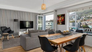 Un salon moderne avec une grande télévision, un canapé gris, une table à manger en bois et des chaises noires.