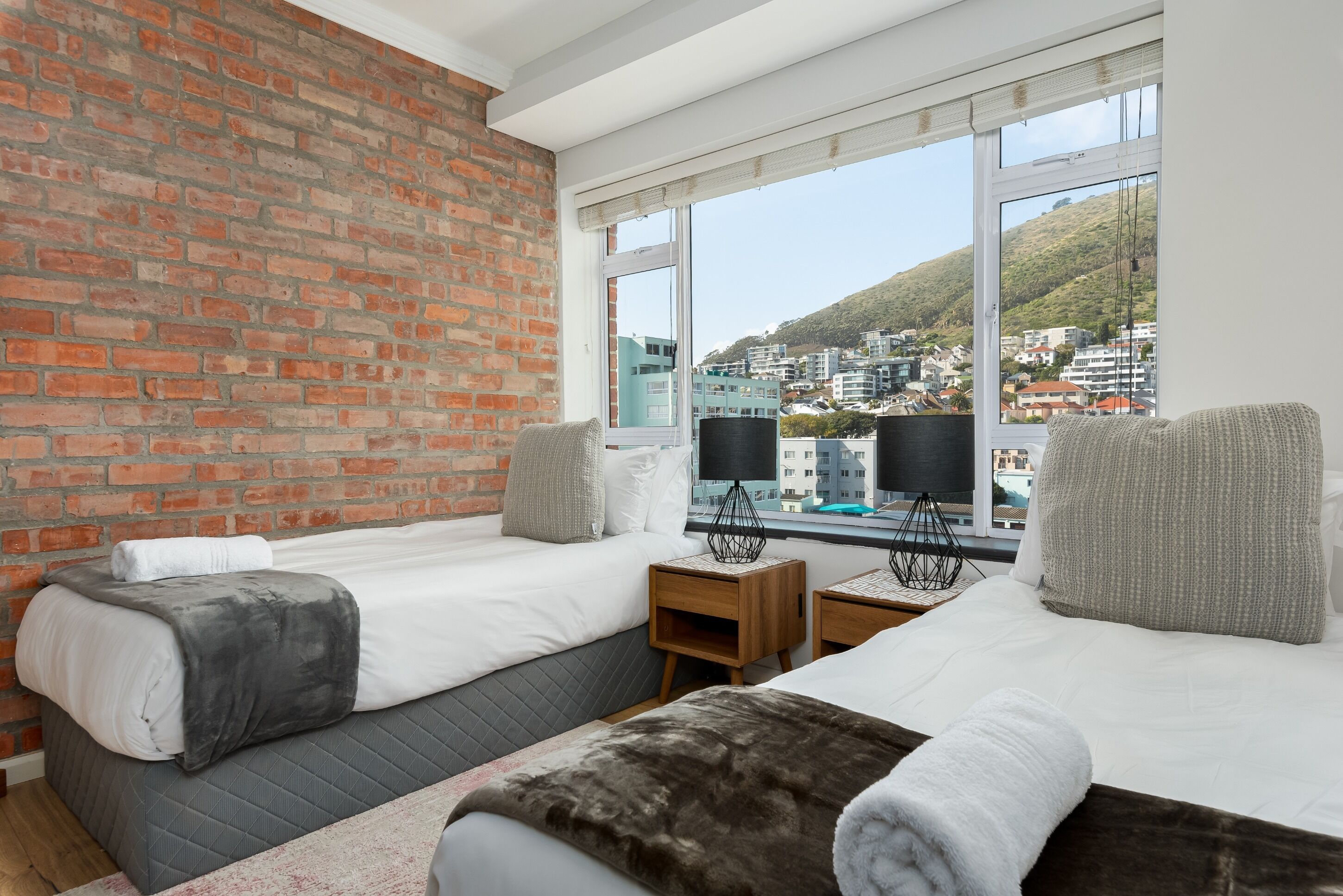 A hotel room with two beds, a brick wall, large windows, and a view of a cityscape.